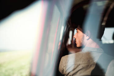 Ein junger hispanischer Mann sitzt in einem Auto auf einem Roadtrip durch die Landschaft, Aufnahme durch Glas, Rückansicht. - HPIF24844