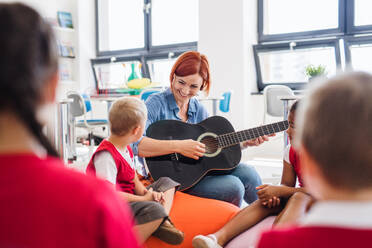 Eine Gruppe von kleinen Schulkindern und ein Lehrer mit Gitarre sitzen in der Klasse auf dem Boden und singen. - HPIF24859