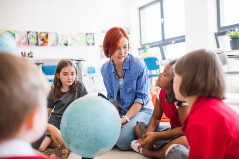 Eine Gruppe kleiner Schulkinder mit einem nicht erkennbaren Lehrer, die in der Klasse auf dem Boden sitzen und Geografie lernen. - HPIF24861
