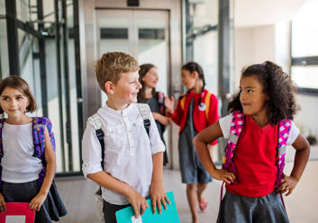 Eine Gruppe von fröhlichen kleinen Schulkindern mit Taschen, die im Korridor spazieren gehen und sich unterhalten. - HPIF24864