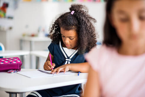 Ein kleines, gemischtrassiges, fröhliches Schulmädchen sitzt am Schreibtisch im Klassenzimmer und schreibt. - HPIF24875