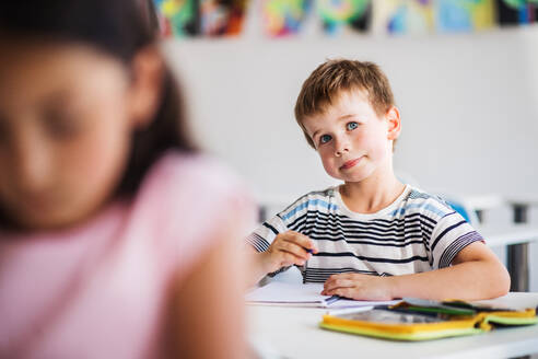 Ein kleiner glücklicher Schuljunge sitzt am Schreibtisch im Klassenzimmer, schreibt und hört dem Lehrer zu. - HPIF24876