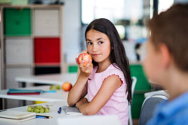 Ein kleines, glückliches Schulmädchen sitzt am Schreibtisch im Klassenzimmer und isst Apfelobst. - HPIF24878