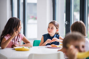 Eine Gruppe fröhlicher kleiner Schulkinder in der Kantine, die zu Mittag essen und sich unterhalten. - HPIF24880