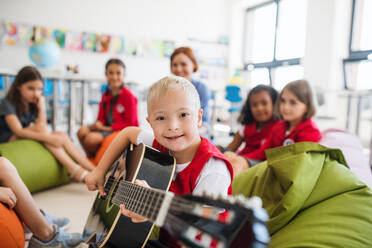 Ein Junge mit Down-Syndrom sitzt mit Schülern und Lehrern in der Klasse auf dem Boden und spielt Gitarre. - HPIF24918