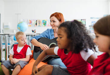 Eine Gruppe von kleinen Schulkindern und ein Lehrer mit Gitarre sitzen in der Klasse auf dem Boden und singen. - HPIF24922