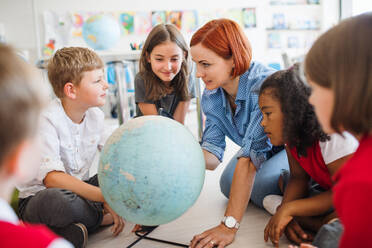 Eine Gruppe kleiner Schulkinder mit einem nicht erkennbaren Lehrer, die in der Klasse auf dem Boden sitzen und Geografie lernen. - HPIF24924