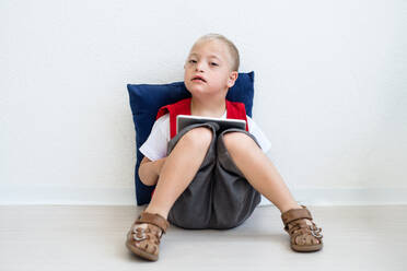 Ein Porträt eines glücklichen Schuljungen mit Down-Syndrom, der auf dem Boden sitzt und ein Tablet benutzt. - HPIF24934