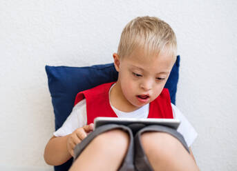 A portrait of happy down-syndrome school boy sitting on the floor, using tablet. - HPIF24936