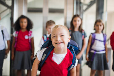 Ein Schuljunge mit Down-Syndrom, der mit einer Tasche und einer Gruppe von Kindern im Korridor spazieren geht. - HPIF24941