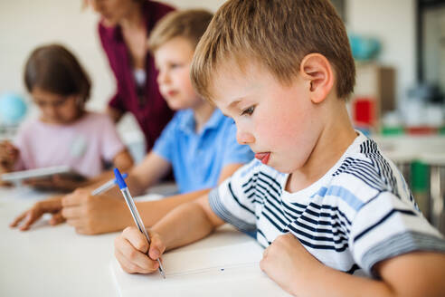 Eine Gruppe von kleinen Schulkindern mit einem Lehrer, der in der Klasse am Schreibtisch sitzt und schreibt. - HPIF24958