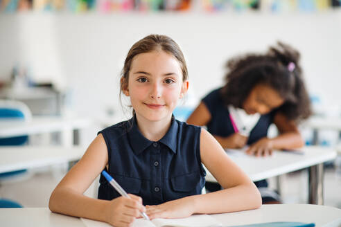 Ein Porträt eines kleinen glücklichen Schulmädchens, das am Schreibtisch im Klassenzimmer sitzt und in die Kamera schaut. - HPIF24968