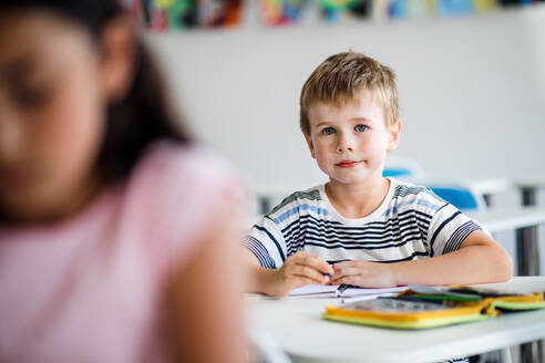 Ein kleiner glücklicher Schuljunge sitzt am Schreibtisch im Klassenzimmer und schaut in die Kamera. - HPIF24971