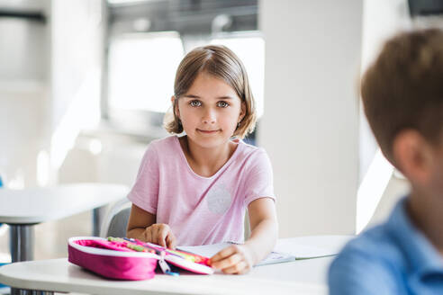 Ein Porträt eines kleinen glücklichen Schulmädchens, das am Schreibtisch im Klassenzimmer sitzt und in die Kamera schaut. - HPIF24972