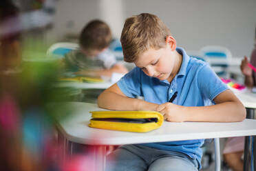 Ein kleiner glücklicher Schuljunge sitzt am Schreibtisch im Klassenzimmer und schreibt Notizen. - HPIF24975