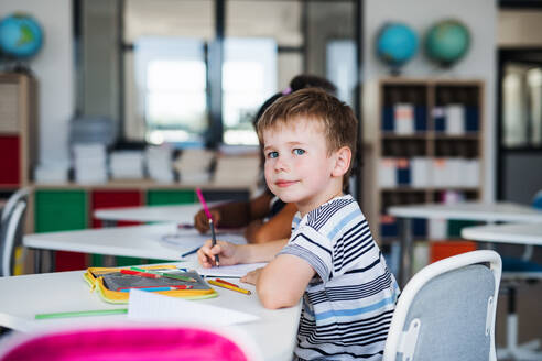 Ein kleiner glücklicher Schuljunge sitzt am Schreibtisch im Klassenzimmer und schreibt Notizen. - HPIF24981