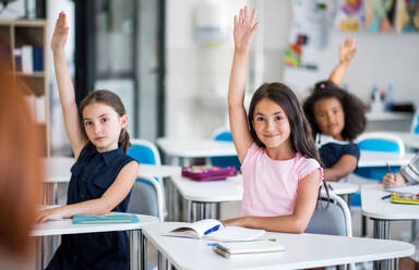 Kleine Schulkinder, die im Klassenzimmer am Schreibtisch sitzen und die Hände heben. - HPIF24983