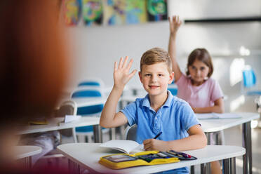 Kleine Schulkinder, die im Klassenzimmer am Schreibtisch sitzen und die Hände heben. - HPIF24984