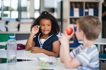 Kleine Schulkinder sitzen am Schreibtisch im Klassenzimmer und essen Obst als Snack. - HPIF24998