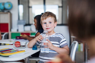 Ein kleiner Schuljunge sitzt am Schreibtisch im Klassenzimmer und trinkt Wasser. - HPIF24999