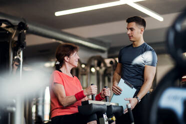 A cheerful female senior with a young trainer doing strength workout exercise in gym. - HPIF25015