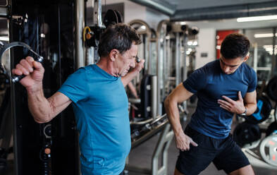 A senior man with a young male trainer doing strength workout exercise in gym. - HPIF25020