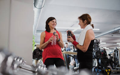 Zwei fröhliche ältere Frauen im Fitnessstudio, die sich nach dem Sport ausruhen und Wasserflaschen in der Hand halten. - HPIF25041