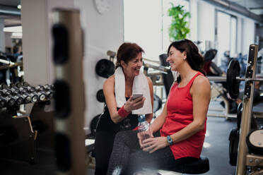 Two cheerful senior women in gym resting after doing exercise, holding smartphone and water bottle. - HPIF25042