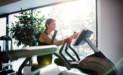 Eine fröhliche Seniorin in einem Fitnessstudio, die ein Ausdauertraining auf einem stationären Fahrrad absolviert. - HPIF25051