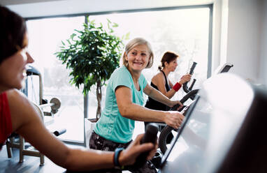 Eine Gruppe von Senioren in einem Fitnessstudio mit einem jungen Trainer, der ein Ausdauertraining auf einem stationären Fahrrad absolviert. - HPIF25052