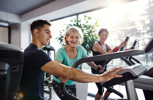 Zwei fröhliche Seniorinnen in einem Fitnessstudio mit einem jungen Trainer, der ein Ausdauertraining auf einem stationären Fahrrad absolviert. - HPIF25053