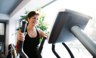 Eine fröhliche Seniorin in einem Fitnessstudio, die ein Ausdauertraining auf einem stationären Fahrrad absolviert. - HPIF25054