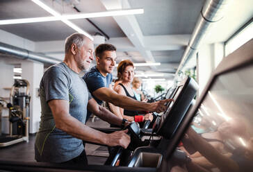 Eine Gruppe von Senioren in einem Fitnessstudio mit einem jungen Trainer, der ein Ausdauertraining auf einem stationären Fahrrad absolviert. - HPIF25057