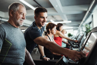 Eine Gruppe von Senioren in einem Fitnessstudio mit einem jungen Trainer, der ein Ausdauertraining auf einem stationären Fahrrad absolviert. - HPIF25058