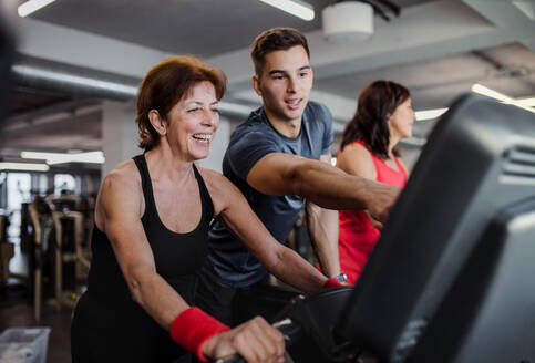 Zwei fröhliche Seniorinnen in einem Fitnessstudio mit einem jungen Trainer, der ein Ausdauertraining auf einem stationären Fahrrad absolviert. - HPIF25059