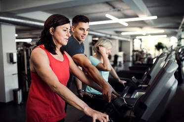 Zwei fröhliche Seniorinnen in einem Fitnessstudio mit einem jungen Trainer, der ein Ausdauertraining auf einem stationären Fahrrad absolviert. - HPIF25060