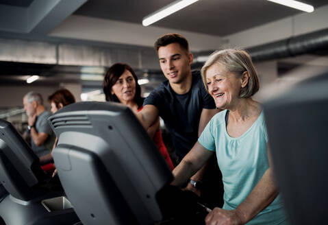Eine Gruppe von Senioren in einem Fitnessstudio mit einem jungen Trainer, der ein Ausdauertraining auf einem stationären Fahrrad absolviert. - HPIF25061
