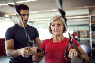 Eine ältere Frau im Fitnessstudio mit einem jungen Personal Trainer bei einer Übung mit TRX. - HPIF25066