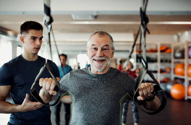 Eine Gruppe fröhlicher Senioren im Fitnessstudio mit einem jungen Personal Trainer, der Übungen mit TRX macht. - HPIF25070