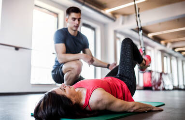 A senior woman in gym and a handsome personal trainer doing exercise with TRX. - HPIF25076
