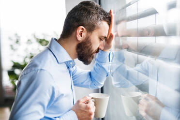 Ein unglücklicher Mann steht mit einer Tasse Kaffee in der Hand am Fenster. - HPIF25235