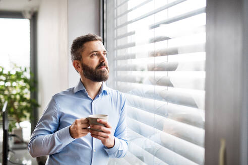 Ein Mann mit einer Tasse Kaffee in der Hand, der am Fenster steht, ein intelligentes Haussteuerungssystem. - HPIF25237