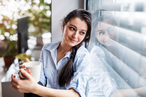 Eine Frau, die mit einer Tasse Kaffee am Fenster sitzt, ein intelligentes Haussteuerungssystem. - HPIF25252