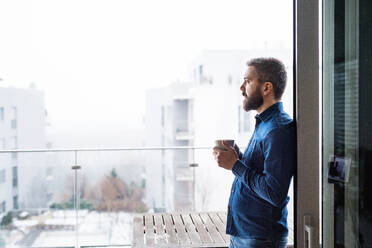 Ein Mann steht mit einer Tasse Kaffee in der Hand zu Hause am Fenster. - HPIF25256