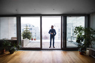 A man standing by the window on a balcony, holding a cup of coffee at home. - HPIF25258