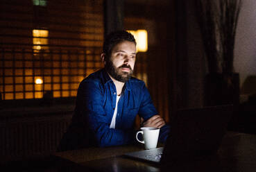 Ein gut aussehender Mann, der zu Hause oder nachts in einem Büro mit einer Tasse Kaffee an einem Laptop arbeitet. - HPIF25266