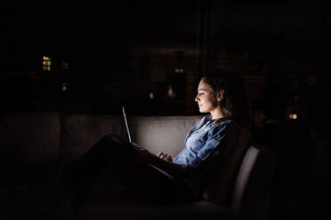 A woman sitting on a sofa at home at night, working on a laptop. - HPIF25273