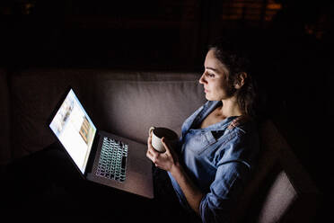 A woman sitting on a sofa at night, working on a laptop. - HPIF25274