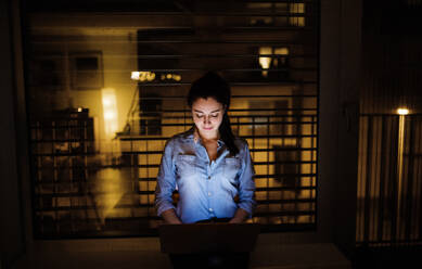 Eine Frau, die nachts auf einer Fensterbank sitzt und zu Hause oder im Büro an einem Laptop arbeitet. - HPIF25276