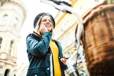 Young beautiful woman with bicycle and smartphone in town, making a phone call. Sunny spring. - HPIF25295